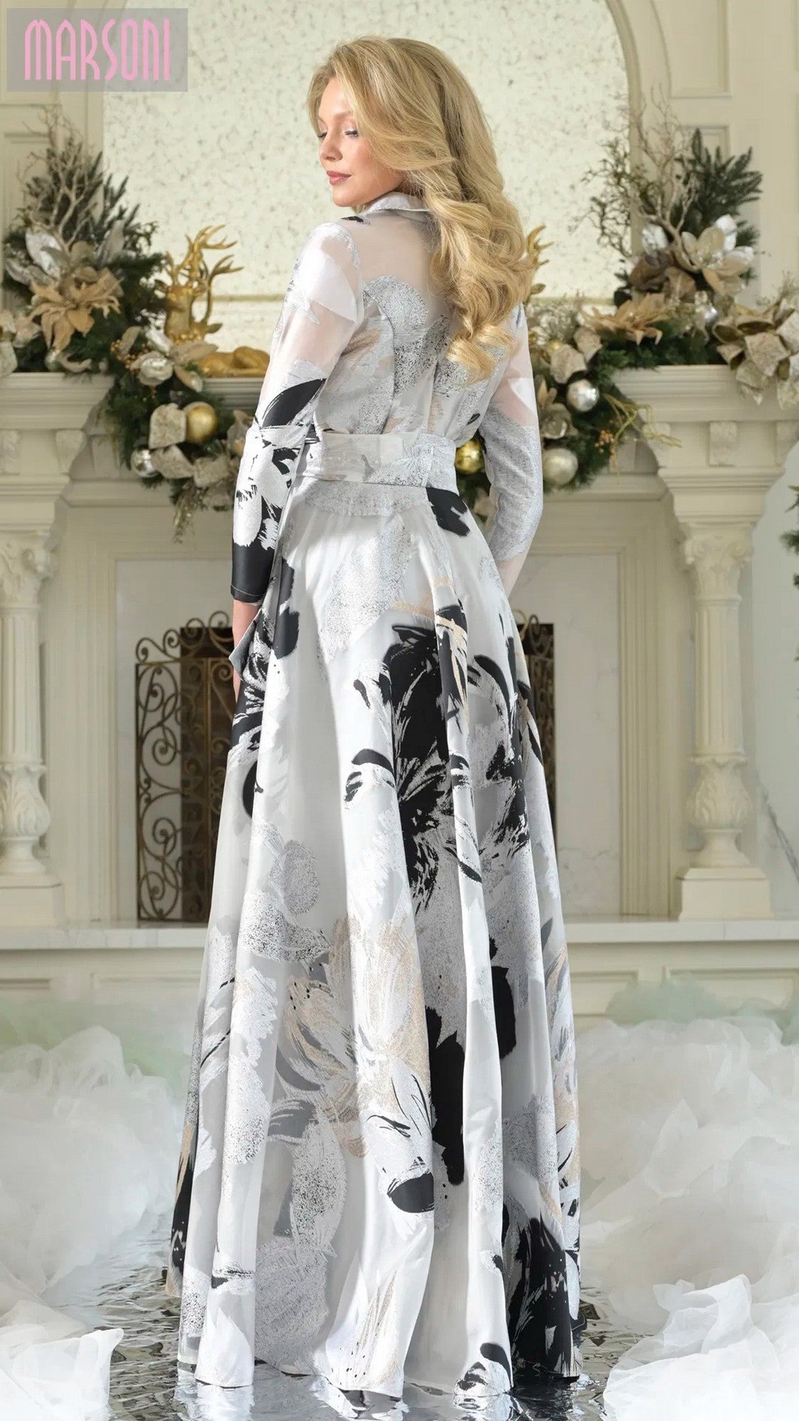 A woman stands indoors in front of a decorated white mantel, wearing the Long Formal Dress: Marsoni MV1367—an organza jacquard, A-line gown adorned with floral and greenery accents in the background.
